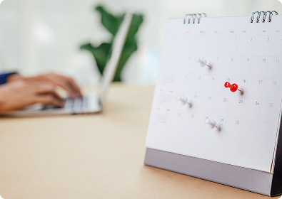 Kalender mit Pinnnadeln auf einem Schreibtisch, Person tippt am Laptop im Hintergrund.